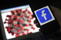 新型コロナウイルスのイラストを背後に、携帯電話の画面に表示されたフェイスブックのロゴ（2020年3月25日撮影）。(c)Olivier DOULIERY / AFP