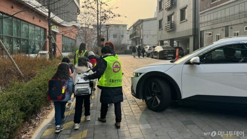 ソウル市東大門区の住宅団地前で、蛍光ベストを着た交通安全指導員が小学校低学年の児童を学校まで引率している(c)MONEYTODAY